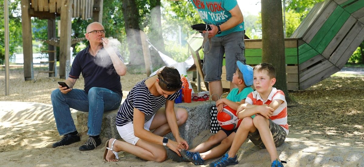 A man vaporizes an e-cigarette on a playground and disturbs a family of mother, father and two kids with his smoke – an appeal for a little more attention and awareness when smoking outdoors. Ein Mann raucht / dampft auf einem Spielplatz eine E-Zigarette und belästigt mit seinem Rauch / Dampf eine Familie mit Mutter, Vater und zwei Jungen – ein Aufruf zu etwas mehr Rücksicht und Achtsamkeit beim Rauchen im Freien.
