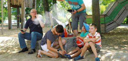 A man vaporizes an e-cigarette on a playground and disturbs a family of mother, father and two kids with his smoke – an appeal for a little more attention and awareness when smoking outdoors. Ein Mann raucht / dampft auf einem Spielplatz eine E-Zigarette und belästigt mit seinem Rauch / Dampf eine Familie mit Mutter, Vater und zwei Jungen – ein Aufruf zu etwas mehr Rücksicht und Achtsamkeit beim Rauchen im Freien.